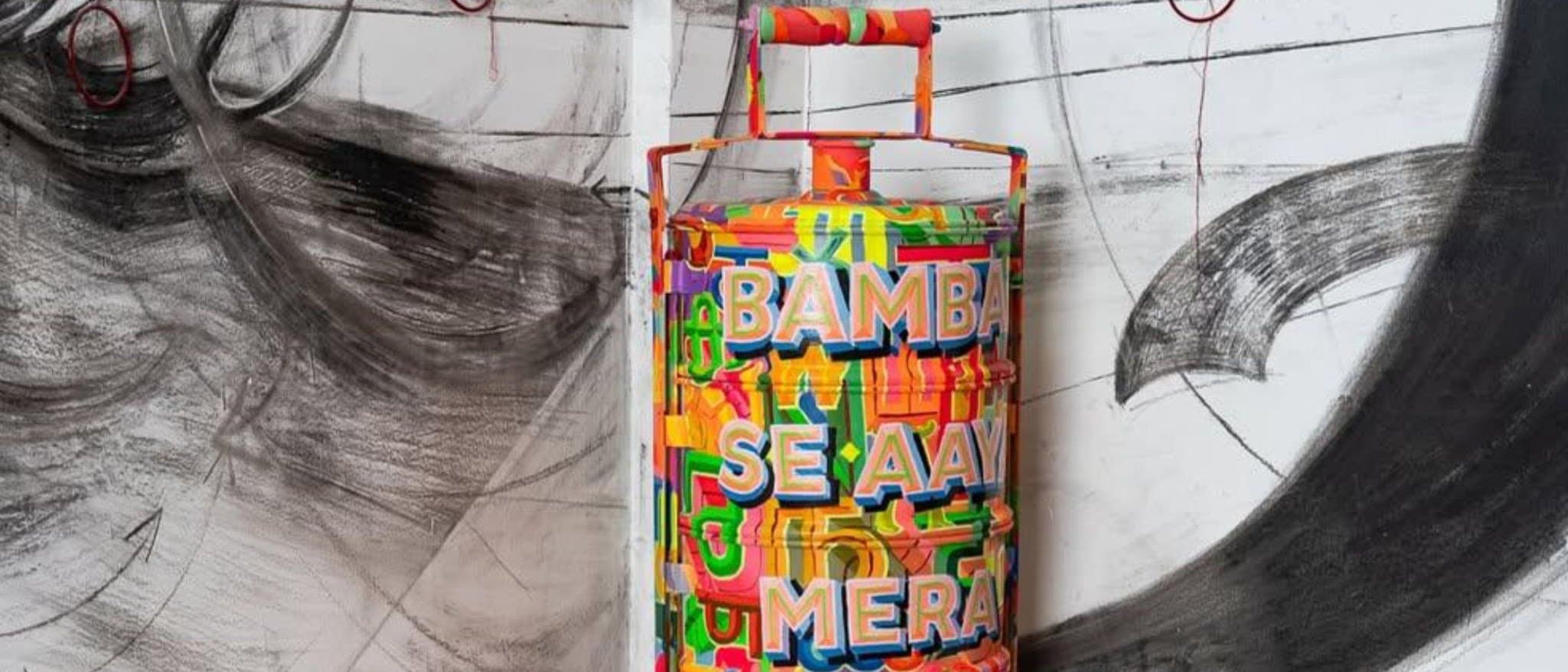 A brightly painted tiffin carrier lettered with the exhibition title, standing in front of a large charcoal wall drawing.