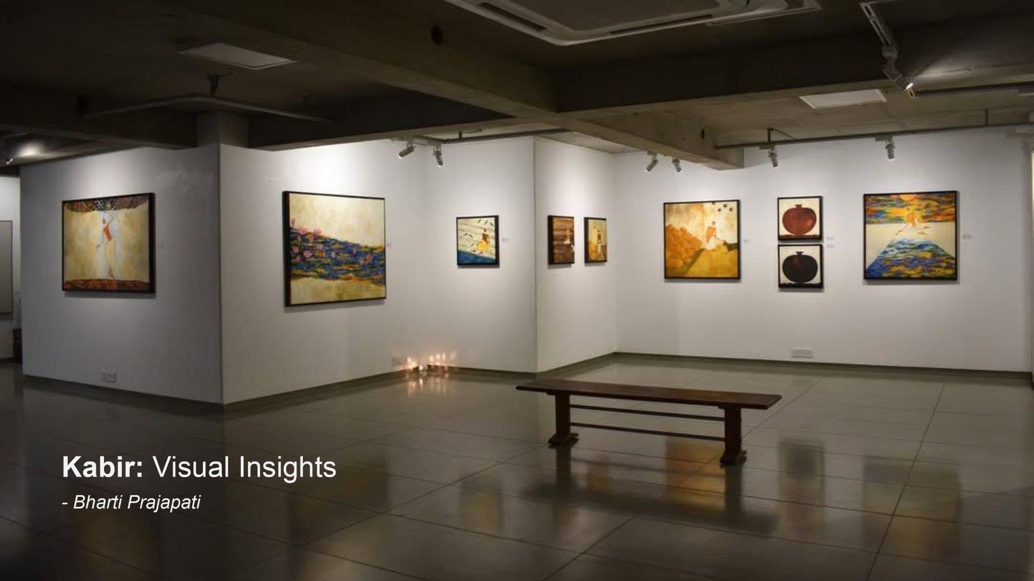 A dimly lit gallery with paintings on white walls, a wooden bench and small lamps on the floor.