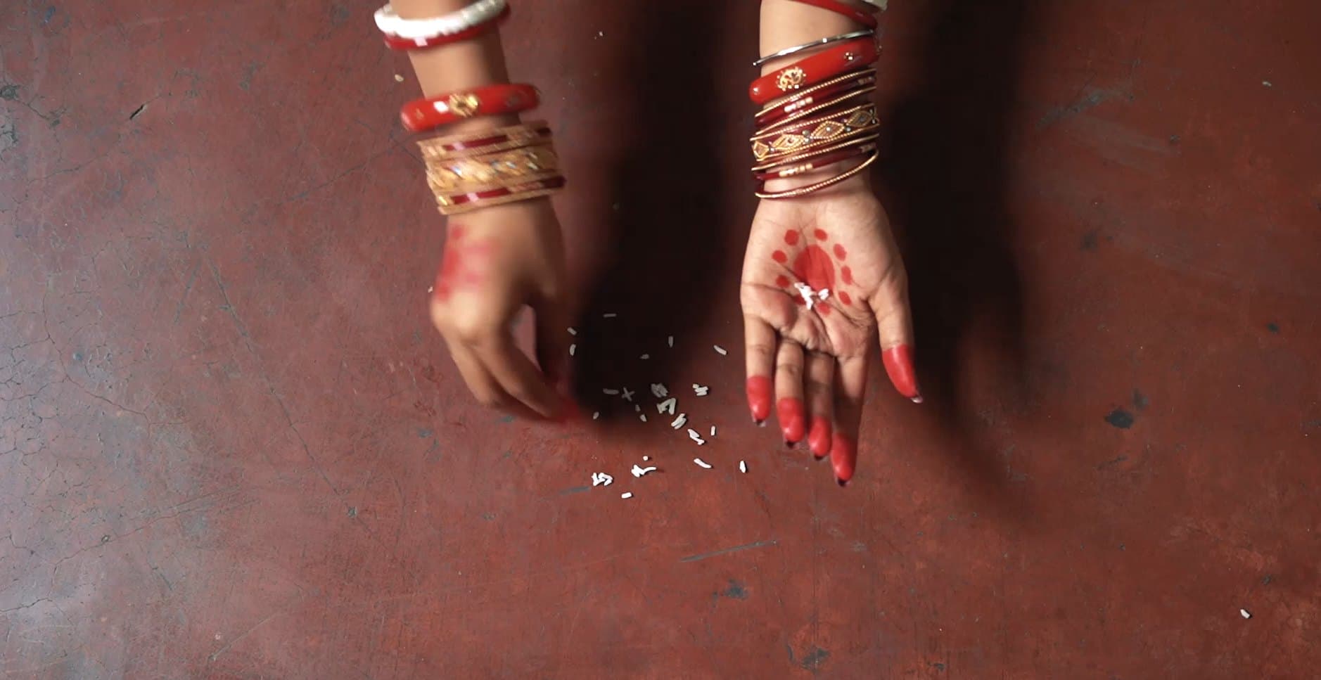 Two hands with red and white bangles and red-stained palms resting on a dark floor, grains of rice scattered between them.